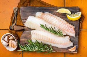 Raw cod fish fillets, codfish with rosemary on wooden board. Orange background. Top view photo