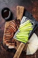 Sliced Peking Duck served with cucumber, green onion, and wheaten chinese pancakes on a meat cleaver. Dark background. Top view photo