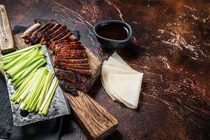 Sliced Peking Duck served with cucumber, green onion, and wheaten chinese pancakes on a meat cleaver. Dark background. Top view. Copy space photo