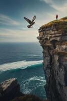 A person stands on a cliff overlooking the ocean with a bird soaring above. photo
