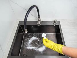 A man's hand in a yellow glove holds a spoon with oxygen bleach against the background of a kitchen sink. Cleaning from grease and dirt. photo
