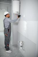 Indian plumber using screwdriver while fixing boiler or water heater, working on heating system in apartment photo