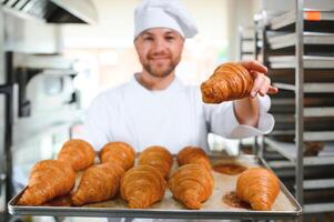 Smiling baker holding tray with fresh cooked croissants in bakehouse photo