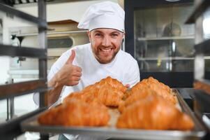 Handsome baker in uniform holding tray full of freshly baked croissants at the manufacturing photo