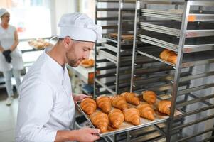 Smiling baker holding tray with fresh cooked croissants in bakehouse photo