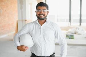Indian construction site manager standing wearing helmet, thinking at construction site. Portrait of mixed race manual worker or architect photo