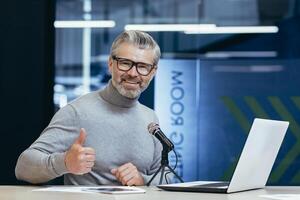 Portrait of a handsome senior man sitting in an office, studio at a table with a microphone. Record podcast, interview, record audio. A smiling man looks at the camera. He points a super finger. photo