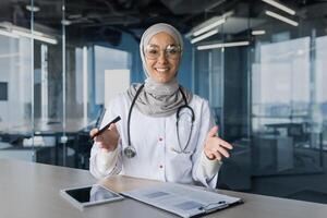 A young beautiful Arab woman, a Muslim doctor is sitting in the office at the desk in a hijab. Conducts a medical online webinar, looks into the camera, explains, smiles. photo