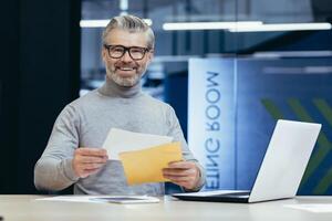 Happy senior man sitting in the office at the desk and holding an envelope in his pocket. I received good news, a loan. I made a contract. He looks at the camera, smiles. photo