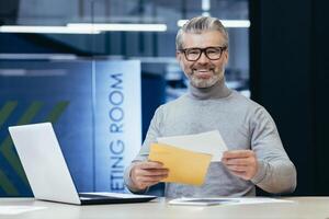 Portrait of successful mature financier, senior gray haired businessman smiling and looking at camera at workplace, boss received notification mail letter, with good news, man working inside office. photo