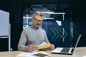 Senior gray-haired man working in office at desk with laptop and documents. He is holding an envelope with a letter, bills, and reports. photo