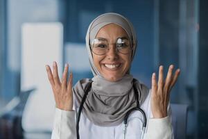 Happy and friendly young beautiful woman doctor student Muslim, Arabic. Standing in hijab, glasses and with stethoscope, looking at camera, pointing with hands. photo