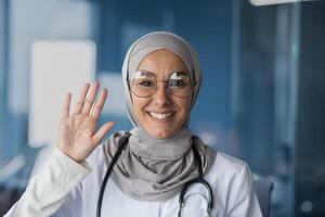 Portrait of a young beautiful woman, a Muslim, Arab woman doctor. She stands dressed in a hijab, glasses and with a stethoscope, looks and waves at the camera, smiles, greets. photo