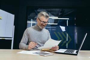Senior gray-haired man working in office at desk with laptop and documents. an envelope with a letter, bills, and reports, good news. photo