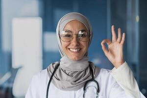 Portrait of a young, beautiful woman, an Arab, Muslim doctor. She stands dressed in a hijab, glasses and with a stethoscope, looks at the camera, smiles, points with her hand ok. photo