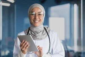 Portrait of a Muslim female doctor in a hijab with a tablet in her hands, the doctor works in the office of a modern clinic, smiles and looks at the camera. photo
