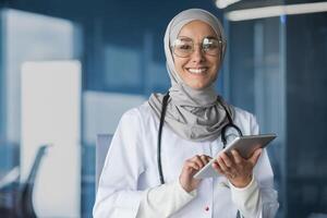 Young beautiful woman asian muslim doctor in hijab smiling and looking at camera working with tablet computer in modern clinic office inside doctor in medical gown and glasses. photo