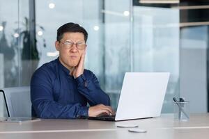 Asian sick in office, businessman has a toothache, man sitting in office inside building working at work using laptop, upset boss in shirt near window. photo