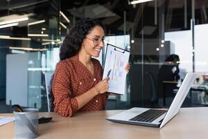Business woman explains report indicators and financial results on call, Hispanic woman smiles and looks at laptop web camera, female worker works inside modern office building with reports. photo