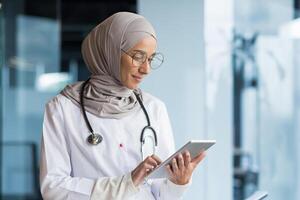 Close-up photo. A young Arab woman, a Muslim doctor is standing in the hospital in the office in a hijab and with a stethoscope. She holds a tablet in her hands, concentrates on working, typing. photo