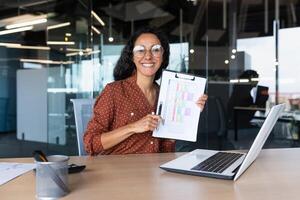Portrait of happy and smiling woman accountant financier, Hispanic woman looking at camera and smiling showing document with financial indicators graphs, businesswoman working inside office. photo