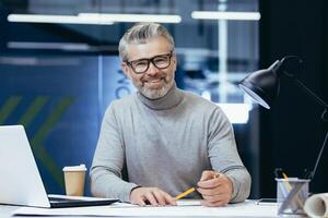 Portrait of an older gray-haired male teacher. He sits at a desk with a laptop, looks at the camera, smiles, holds a pencil in his hands. Teaches online, remotely. photo