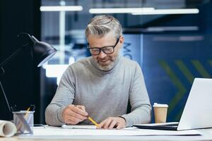 Successful senior architect at work in office, mature gray haired man drawing blueprints on layout and plan smiling and happy with work. photo