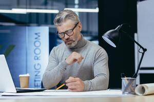 Concentrated senior male architect working in the evening by a lamp in the office. Sitting at the table, drawing a plan with a pencil, working with papers. photo