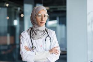 Portrait of a young Muslim woman doctor. She is standing in the hall of the hospital dressed in a hijab, wearing glasses and a stethoscope, looking seriously into the camera, her arms crossed. photo
