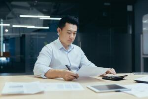 Worried and serious young Asian businessman working in office with documents, busy with paper work, checking accounts on calculator. photo