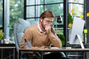 A man in the office at the workplace is sick, he has a severe stomach ache, the businessman got food poisoning from lunch, he is sitting at the table inside the building. photo