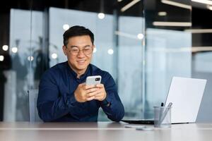 Cheerful and smiling asian businessman inside office using phone, man typing message and browsing online pages and watching , boss in glasses and shirt at work. photo