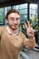 Man in office wearing shirt and glasses smiling and looking at smartphone camera, businessman taking selfie and talking on call using smartphone, programmer waving hand greeting gesture photo