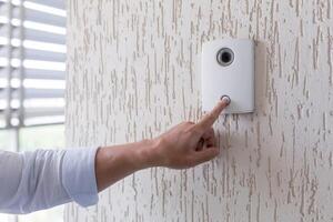Hand of a man in a white shirt pressing the button of a door knocker on a white wall of a house, a smart door knocker with a camera for communication photo