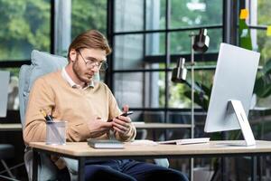 A young programmer, freelancer sits at the desk in the office, works at the computer. Holds a phone in his hands, uses a gadget. Writes a message, checks mail. photo