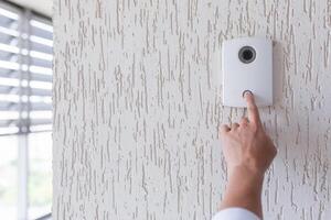 Hand of a man in a white shirt pressing the button of a door knocker on a white wall of a house, a smart door knocker with a camera for communication photo