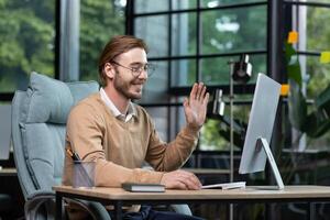 Successful, satisfied young boss conducts an online call with colleagues, sits at the table, looks at the computer monitor, waves at the webcam in greeting. photo