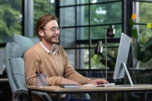 Young successful entrepreneur working inside office with computer, man in casual clothes smiling and typing on keyboard, businessman happy with achievement results and income. photo