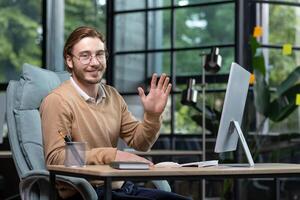 Portrait of a young businessman, designer, programmer in a busy office sitting at a table with a computer, smiling at the camera, waving, greeting, introducing himself. photo