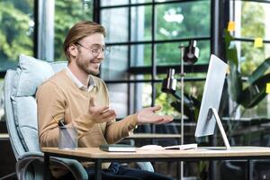 Happy and successful man at work looking at computer web camera smiling and talking with colleagues on call, businessman working inside office building, remote online meeting with friends photo