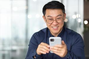 Close-up photo of a happy and smiling Asian man reading happy news from the phone, a businessman in a shirt and glasses smiling and rejoicing, a man works inside the office near the window.
