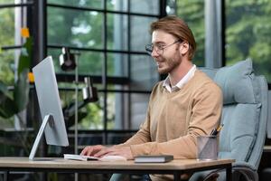 A young male programmer, freelancer sits in the office at the table near the window and works at the computer, typing on the keyboard. Smiling, wearing glasses and a beige sweater. photo