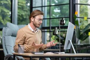 An angry young man sits at the table, looks at the computer screen, spreads his hands, does not understand what the problem is, non-working device, bad internet, call. photo