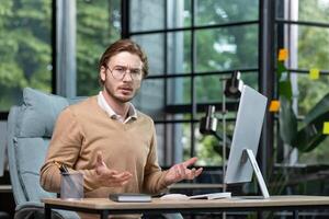 Solving a problem, making a decision. Portrait of a young freelancer, a programmer who looks worriedly and angrily at the camera, spreads his hands. Sitting in the office at the desk. photo