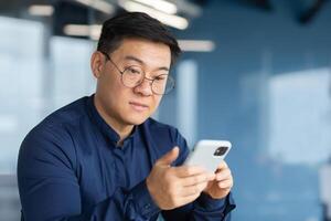 Close-up of serious and pensive Asian businessman using phone, man inside office typing message, browsing internet pages and watching . photo