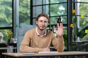 Portrait of a young businessman sitting at a desk in the office, waving his hand in greeting, looking at the camera with a serious expression on his face. photo