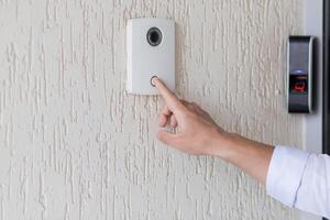 Hand of a man in a white shirt pressing the button of a door knocker on a white wall of a house, a smart door knocker with a camera for communication photo
