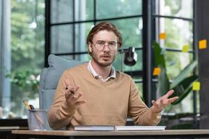Online business meeting. Serious young man businessman, programmer sits in the office and stares at the camera. Confused communicates with clients, partners. Gesturing with hands. photo