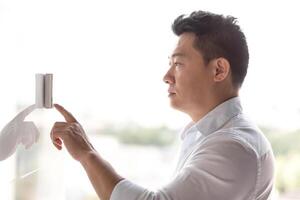An Asian man stands near an electronic panel on the wall and presses the entry bell of an automatic door with his finger. Configures the wall conditioner, the security system. photo