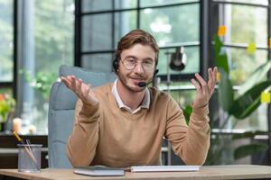 Portrait of a young male blogger in a headset sitting at a desk in the office with a camera and talking via call, recording a podcast, developing his online channel. photo
